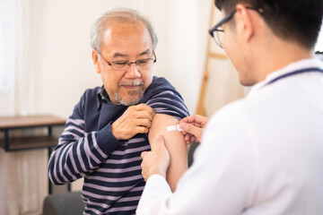 Safe vaccination for old people. Elder man in medical  getting flu or Covid-19 vaccine sitting on sofa at home. Asian Doctor or nurse giving flu or Covid-19 shot to senior. - obrazy, fototapety, plakaty