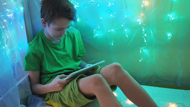 a boy is playing a tablet sitting on a bed decorated with garlands. multicolored light of lights. a teenager in a green T-shirt and shorts