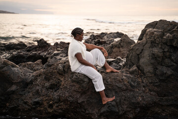 Beautiful pregnant woman in white clothes posing on the rocky coast, sitting and relaxing during sunset. Future motherhood concept. Mom to be.