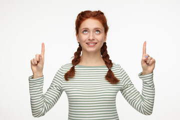 Indoor portrait of young ginger female posing over white background point with a finger above at copy space