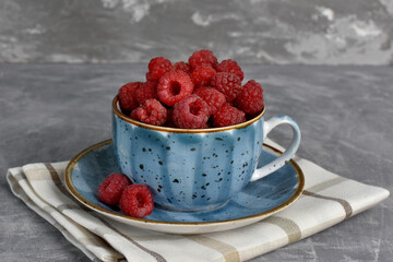 ripe red raspberries in a beautiful blue cup on a gray stone background. summer berries
