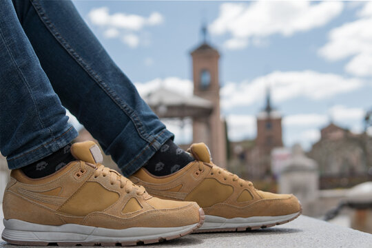 Close shot of a pair of brown leather sneakers with a city background behind on a sunny day. Legs of person in jeans and khaki or yellow shoes stepping on the platform and with copy space.
