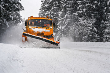 Orange maintenance plough truck on forest road after snowstorm blizzard. Roads get dangerous during winter (driver face blurred) - obrazy, fototapety, plakaty