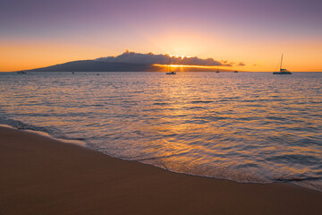 Rays of sunset illuminate peaceful scene of clouds, sky, sea and sailboats anchored off a sandy...