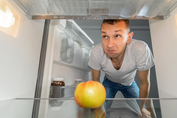 A disgruntled, hungry man is looking for food in the refrigerator and looks in surprise at an apple core in an empty refrigerator. Photo from inside the refrigerator