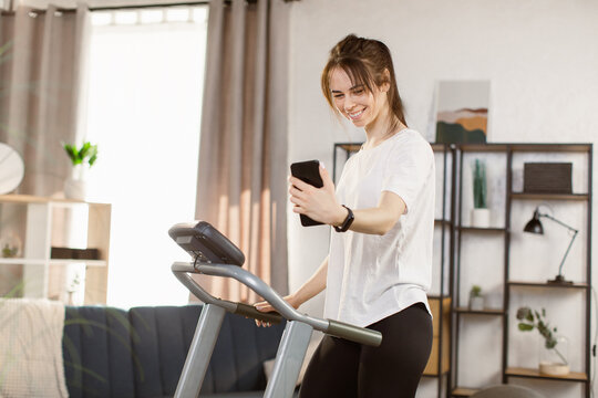 Young woman doing morning exercises and speaking on the phone at home. Female athlete workout running on treadmill at modern apartment. Concept of training in modern gym and pump up body