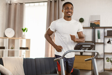 Fototapeta na wymiar Side view of african american sports man in sports shorts and a white t-shirt doing cardio training on treadmill at home looking away. Concept of sport, health, action, nutrition.