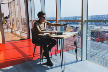 Young handsome smiling smart freelancer in glasses works at a laptop while spending time in a cafe. A positive student sits at a wooden table in a cafe. Happy man sitting while working indoors