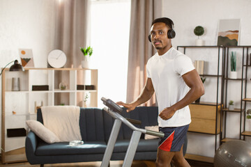 Fototapeta na wymiar Young african man jogging on the modern treadmill listening to music on headphones in shorts and a white t-shirt running on a treadmill at home. Male runner doing cardio training.