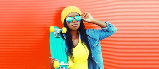 Rolgordijnen Portrait of young african woman with skateboard wearing sunglasses, hat in casual on vivid background, blank copy space for advertising text © rohappy