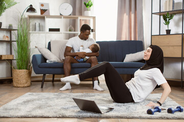 An Arabian muslim woman in a sports leggings and a white T-shirt is practicing yoga in the livingroom with her african american husband and son on the background.