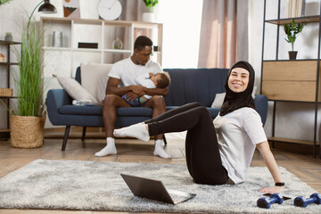 Obraz premium A girl in a hijab is engaged in yoga at home. Muslim woman working out at home online. Girl in sportswear on a floor mat doing abs exercises with her husband and son on the background.