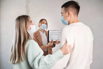 Fototapeta premium Woman in suit talking to couple and show apartment plan in unfinished building. All people in masks. Life in pandemic