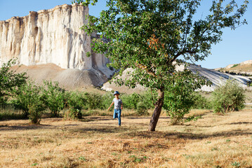 A cheerful kid in jeans, a T-shirt and a blue hat is playing in the garden among the apple trees.