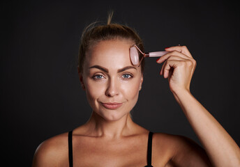 Obraz premium Attractive beautiful woman with tanned healthy glowing skin massages her face with jade stone roller massager, performs lymphatic drainage massage for skin smoothing and anti-wrinkles beauty treatment