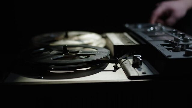 close up old reel-to-reel tape recorder plays magnetic tape record. Hand push start button. Beautiful gloomy shot of vintage retro turntable on table dark light. beam of light falls on old reel player