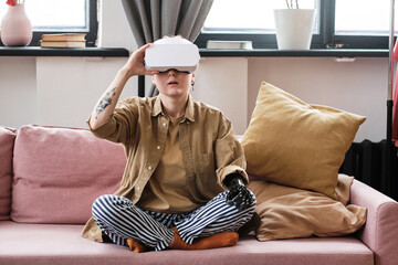 Young woman with arm prosthesis sitting on sofa in living room and using virtual reality headset