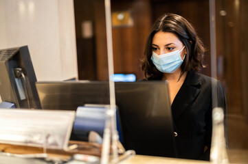 Happy female receptionist standing at hotel counter wearing masks to protect from covid 19...