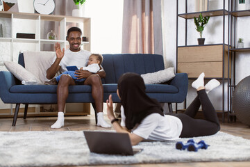 Obraz premium Young sporty african man sitting on sofa holding his multinational son in his arms and waving his hand to muslim arab wife doing working out sports lying on the carpet.