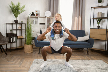 Small son sit on strong dad shoulders doing squat exercises. African family enjoy activity games at home, healthy fit lifestyle, two superheroes, happy Father Day concept