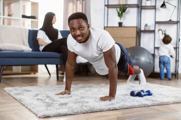 Training At Home. Sporty african man doing yoga plank looking at camera with muslim wife and son on background, exercising in living room, free space