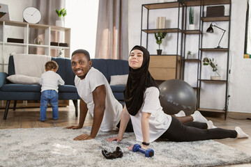 Obraz premium 25-30 age beautiful sporty couple practicing yoga doing four-limbed staff pose at home. Muscular Athletic African Man and Beautiful Muslim Fitness Woman in Workout Clothes Doing Push Up Exercises.