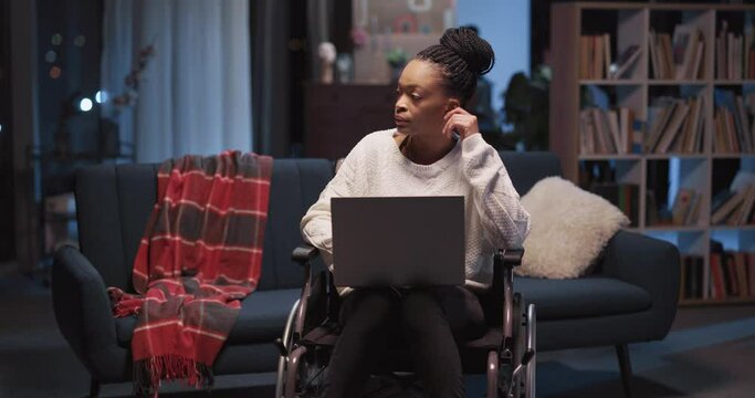 Young beautiful afro-american woman disabled in wheelchair doing freelance work project in the living room. Workaholic girl. Disabilities. Healthcare.