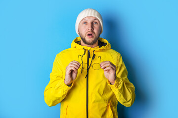 Blind young man in a yellow jacket holding glasses in his hands on a blue background