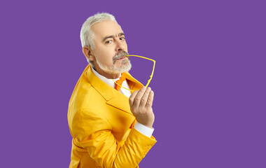 Portrait of confident handsome senior man in funky yellow suit holding his glasses and looking at...