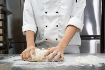 Selective focus of two hands of an Asian male baker in a white chef dress, standing kneading bread dough on a stainless steel table full of flour in front of a blurred refrigerator in a bakery kitchen