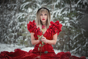 Young woman in the red dress in the snowy winter forest with red rose flower.