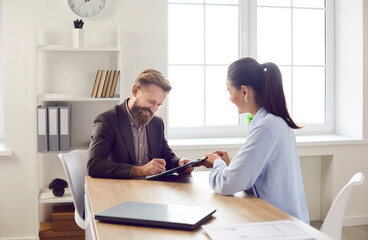 Happy client signing some paper documents. Smiling bearded man in suit sitting at office desk with...