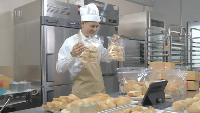 A middle-aged Asian male baker in a white chef uniform standing at a table showing the delicious bread in transparent bags in front of the tablet, doing a live selling online of bread in a kitchen.