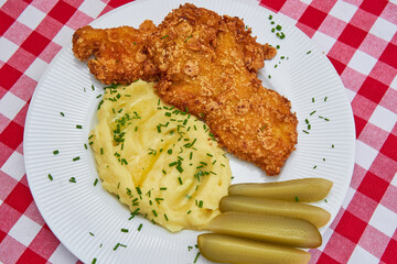 Close up picture of deep fried chicken schnitzel with mashed potatoes and pickled sour cucumbers sprinkled with minced chives served on the white rustic plate. Traditional dish from Czech Republic. 