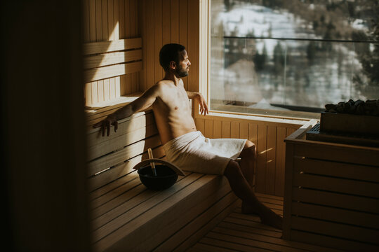 Young man relaxing in the sauna and watching winter forest through the window