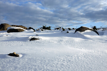 Winter tundra landscape with hilly snowy surface with stones and large boulders and long shadows from them under a beautiful blue sky with clouds