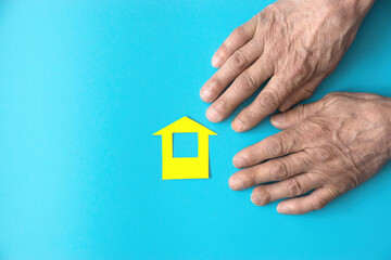 Hands of an elderly man and a yellow paper-cut house on a blue background. Topic homeless, nursing home, loneliness in old age. Mortgage concept, real estate purchase.