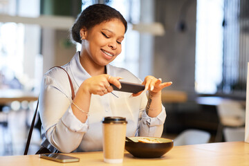 Portrait of young black woman taking photo of food with smartphone while sitting at table in Asian...