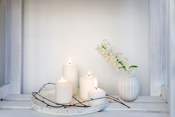 Fototapeta na wymiar A bouquet of white cut hyacinth a in a small white corrugated vase and three large burning candles on a round tray are on a beige table. Place for text