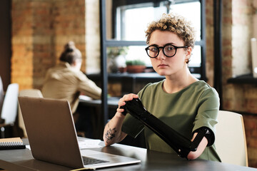 Portrait of young woman in eyeglasses with prosthetic arm sitting at her workplace with laptop and...