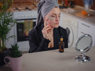 Sculpting, lifting and anti-aging care. Skin care and beauty at home. Woman using calming quartz roller massager while sitting on sofa in kitchen looking in mirror. Woman using face roller.