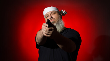 A fat man with a gray beard and a bandage on his head aims with a gun on a dark background