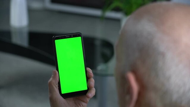 Bald hoary elderly man holds smart phone with empty green screen for mockup