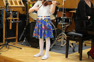 A little girl with a violin standing up plays a musical instrument a piano teacher trains a young student at a school concert a kid on stage with equipment drums and a microphone