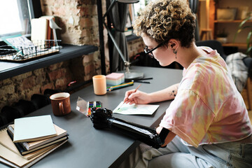 Girl with prosthetic arm sitting at table and painting a picture during her leisure time