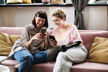 Happy young woman looking at mobile phone and smiling while sitting on sofa with her friend with...