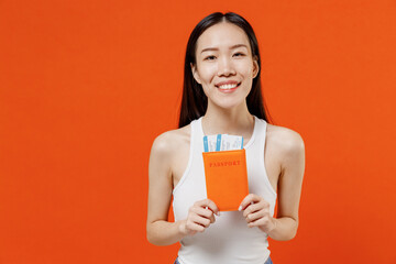Smiling fun excited jubilant exultant young woman of Asian ethnicity 20s years old in white tank top hold passport boarding tickets looking camera isolated on plain orange background studio portrait.