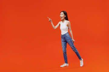 Full size body length side view impressed fun young woman of Asian ethnicity 20s in white tank top pointing index finger aside keep mouth wide open isolated on plain orange background studio portrait. - obrazy, fototapety, plakaty