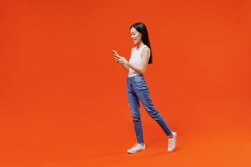 Fototapeta na wymiar Full size side view happy young woman of Asian ethnicity 20s year old in white tank top hold in hand use mobile cell phone typing browsing chatting isolated on plain orange background studio portrait