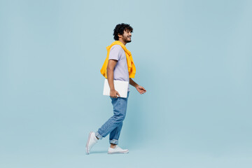 Full size body length side view exultant young bearded Indian man 20s wears white t-shirt hold use work on laptop pc computer stepping isolated on plain pastel light blue background studio portrait.